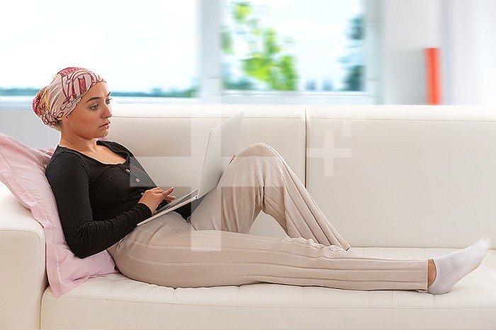 Woman with a scarf in her hair lying on the sofa, a laptop on her knees.