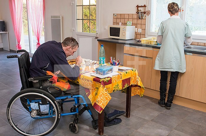 ADMR 62 - Home Help in Rural Areas, Pas de Calais. Muriel, Auxiliary of Social Life (AVS) works at the home of Mr. T. who is severely handicapped by Multiple Sclerosis. Muriel must help her with all the daily tasks. Disabled person in a wheelchair at the table eating and his carer doing the dishes.