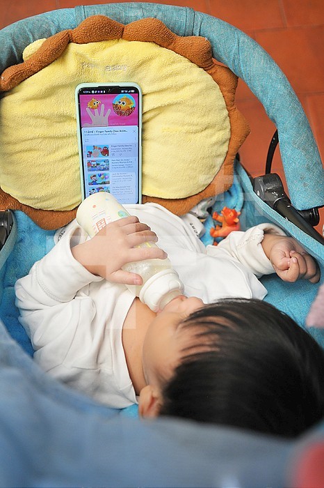 Young child in his cradle with his bottle in front of a phone screen.